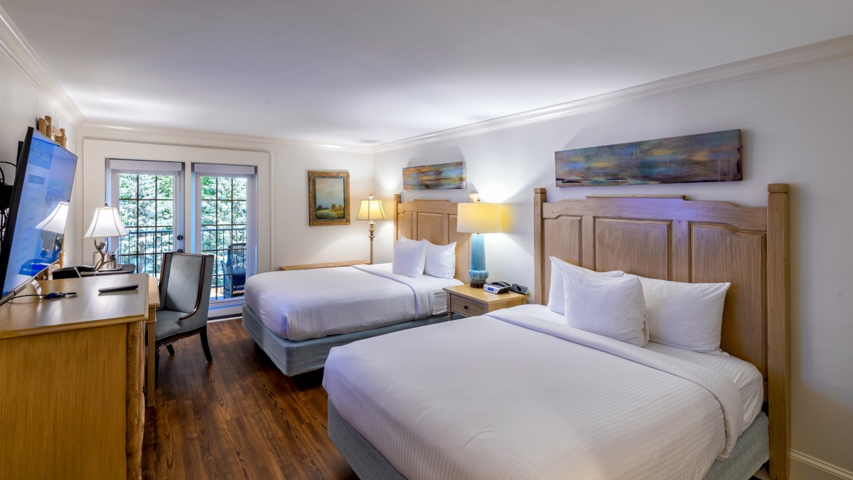 The image shows a hotel room with two beds, lamps, artworks, a desk, and a TV, with natural light coming through French doors.