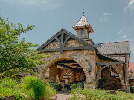A rustic building with stone arches and a metal roof, surrounded by greenery and under a partly cloudy sky.
