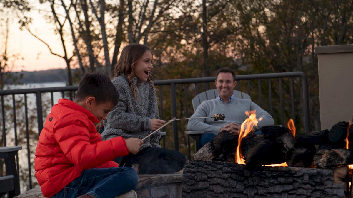A group of people enjoying a campfire on a patio at sunset, two kids roasting marshmallows while a man watches.