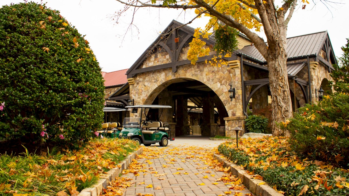 A stone building with an arched entrance is surrounded by autumn leaves, trees, and golf carts on a brick path.