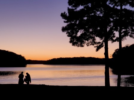 The image shows a scenic sunset over a lake with silhouettes of two people and trees in the foreground.