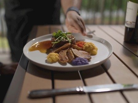 A hand serves a gourmet dish with meat, colorful mashed potatoes, and garnish on a table beside wine and flowers.