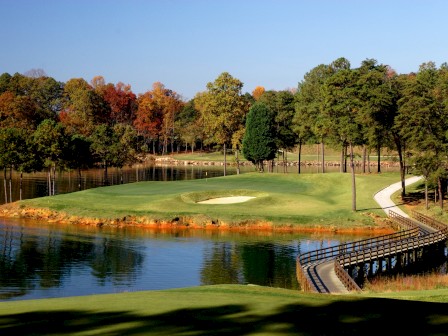 A golf course with a bridge over water, surrounded by trees and a pathway leading to the green under a clear blue sky.