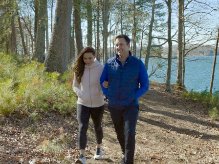 A couple is walking on a wooded path near a lake, surrounded by trees and sunlight filtering through the branches.