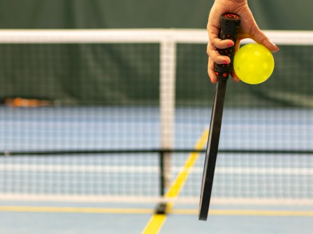 A person holds a paddle and a yellow ball on a court with a net in the background, likely related to pickleball.