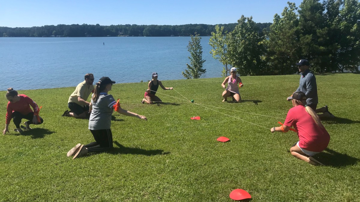 People are kneeling in a circle on grass near a lake, playing a game with cones in the sunlight.