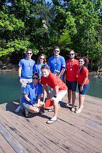 A group of people wearing blue and red shirts standing on a wooden dock by a body of water, with trees in the background.