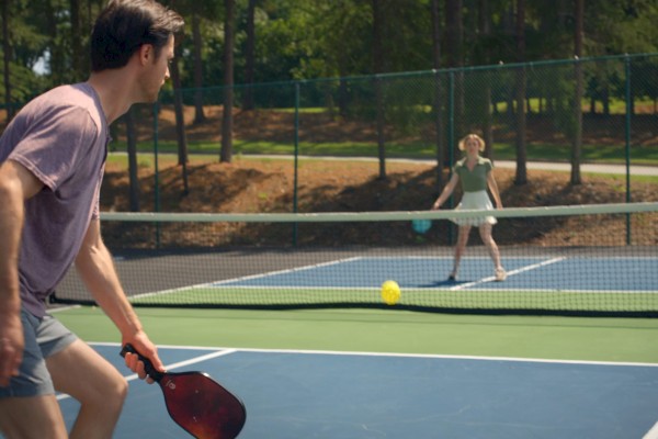 Two people are playing pickleball on an outdoor court, with a yellow ball between them.