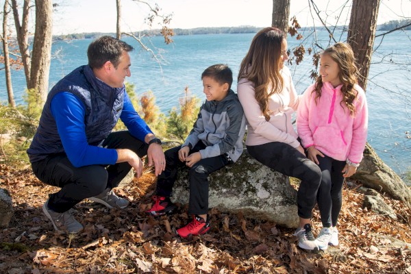 A family sits outdoors by a lake, surrounded by trees. They're dressed warmly, with the dad squatting beside the mom and two kids on a rock.