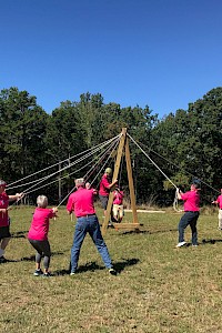 People in pink shirts are playing with a maypole in an open grassy area with trees in the background.