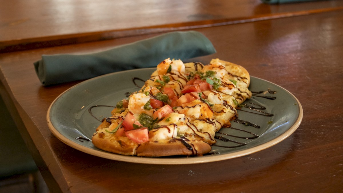 A flatbread topped with cheese, tomatoes, greens, and a drizzle of balsamic glaze sits on a plate on a wooden table.