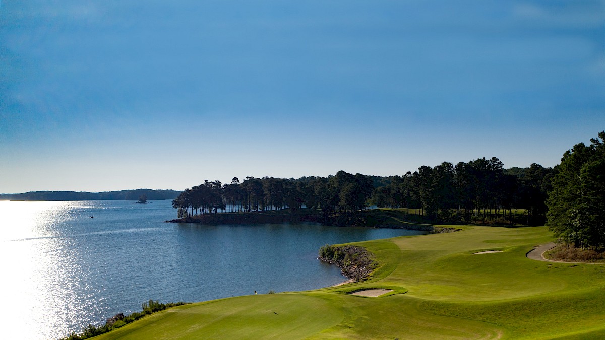 A golf course by a serene lake, lined with trees under a clear blue sky, reflecting sunlight on the water.