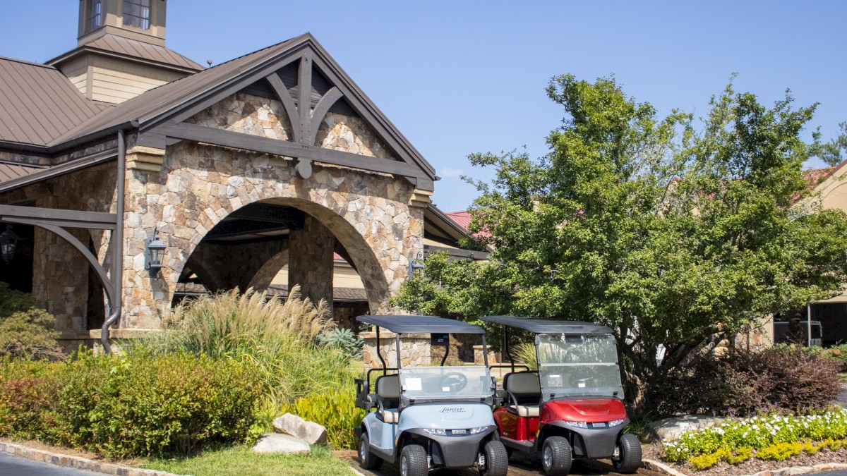 The image shows two golf carts parked in front of a stone building with an archway, surrounded by greenery and a clear blue sky.