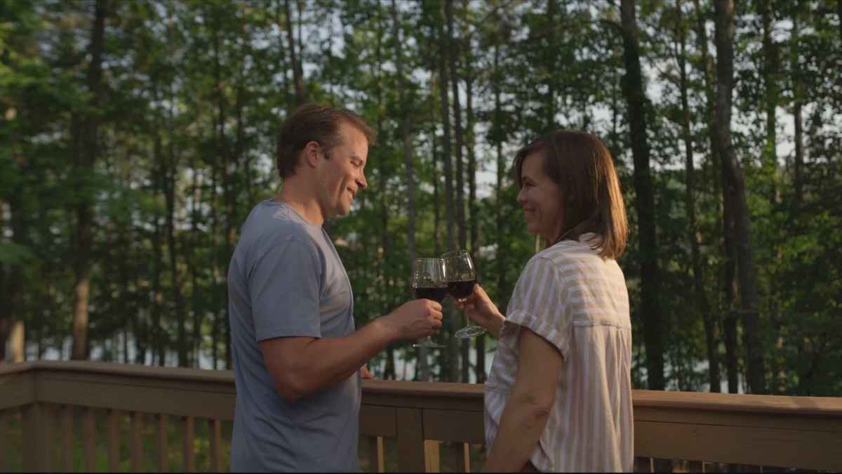 A couple is on a deck surrounded by trees, holding glasses of red wine, and smiling at each other in the evening light.