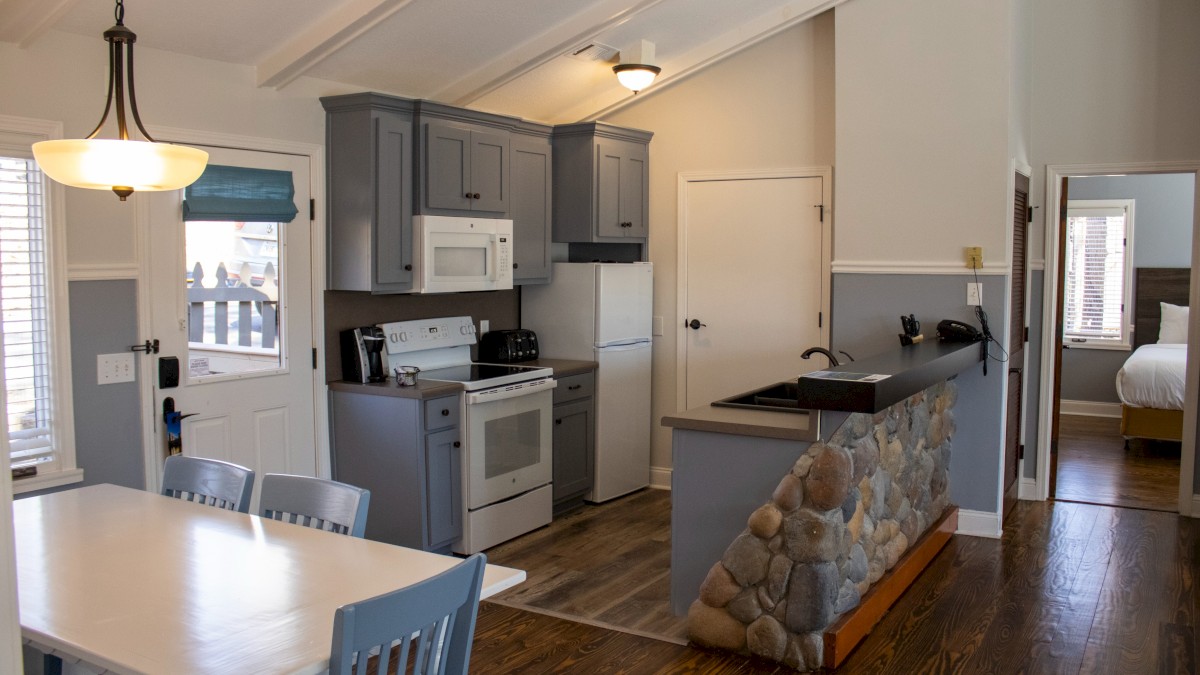 A cozy kitchen with gray cabinets, a stone-faced bar, and a dining area, leading to a bedroom with a visible bed.