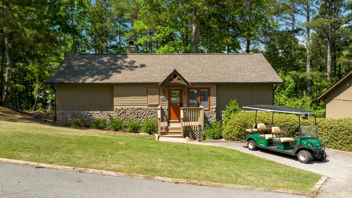 A small house with a sloped roof surrounded by trees, and a green golf cart parked in the driveway on a sunny day.