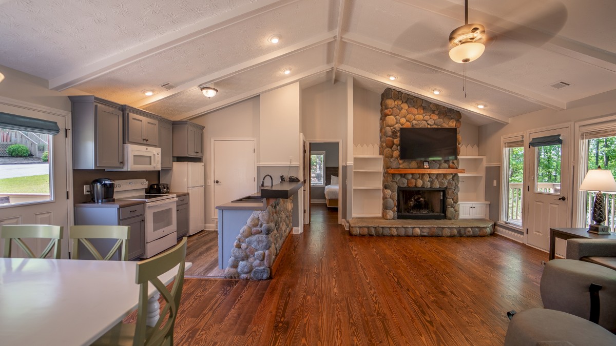 A cozy living room and kitchen area with wooden floors, stone fireplace, dining table, and large windows with forest views.