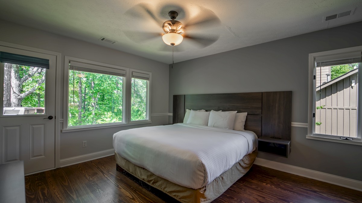 A bedroom with a large bed, wood floor, and ceiling fan, surrounded by windows showing outdoor greenery and a door to the left.