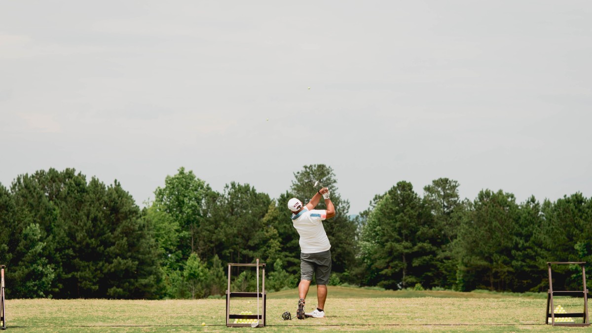 A person is swinging a golf club on a grassy field, surrounded by trees. Golf equipment is visible nearby.