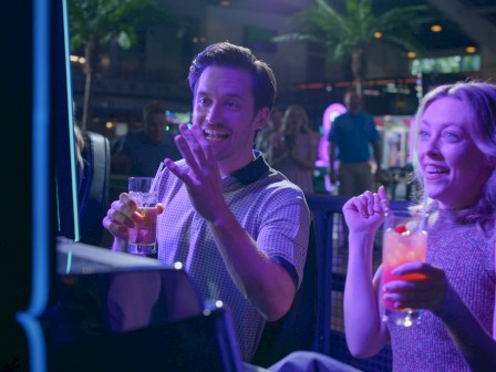 Two people are excitedly playing a game at an arcade, holding drinks and enjoying the vibrant atmosphere.