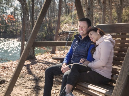 A couple sits on a wooden swing by a lake, surrounded by trees, enjoying a peaceful moment in nature.