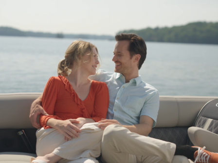 A couple is smiling and relaxing on a boat, with water and distant land visible in the background.