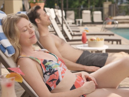 Two people are lounging by a pool on deck chairs, with drinks and snacks nearby, enjoying a sunny day.