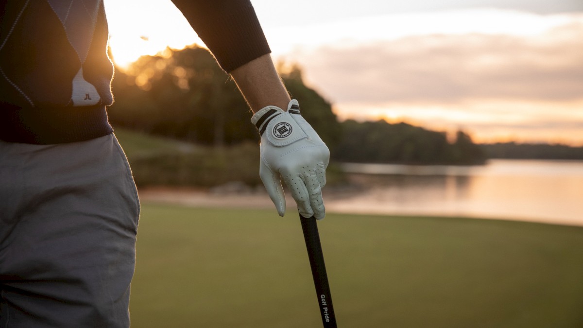 A person stands outdoors on a golf course at sunset, holding a golf club, wearing a glove, with scenic water and trees in the background.