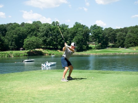A person is playing golf near a lake, preparing to swing. Trees and boats are visible in the background under a clear sky.