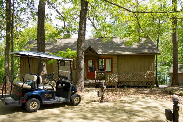 A cabin in the woods with a golf cart parked in front, surrounded by trees and dappled sunlight.