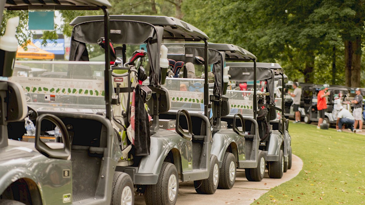 Several golf carts lined up on a path with golf bags, set against a background of trees and a grassy area.