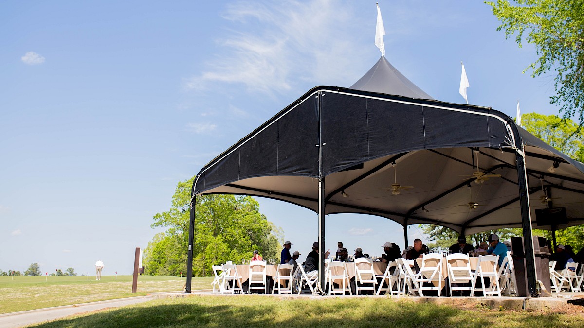 A group of people seated under a large outdoor tent with white chairs, set in a grassy area on a sunny day.
