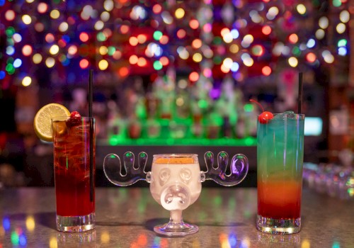 Colorful, festive cocktails with umbrellas and garnishes sit on a bar counter against a blurred, vibrant, and bokeh-lit background.
