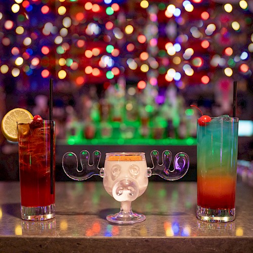 Colorful, festive cocktails with umbrellas and garnishes sit on a bar counter against a blurred, vibrant, and bokeh-lit background.