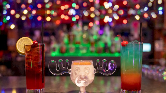 Colorful, festive cocktails with umbrellas and garnishes sit on a bar counter against a blurred, vibrant, and bokeh-lit background.