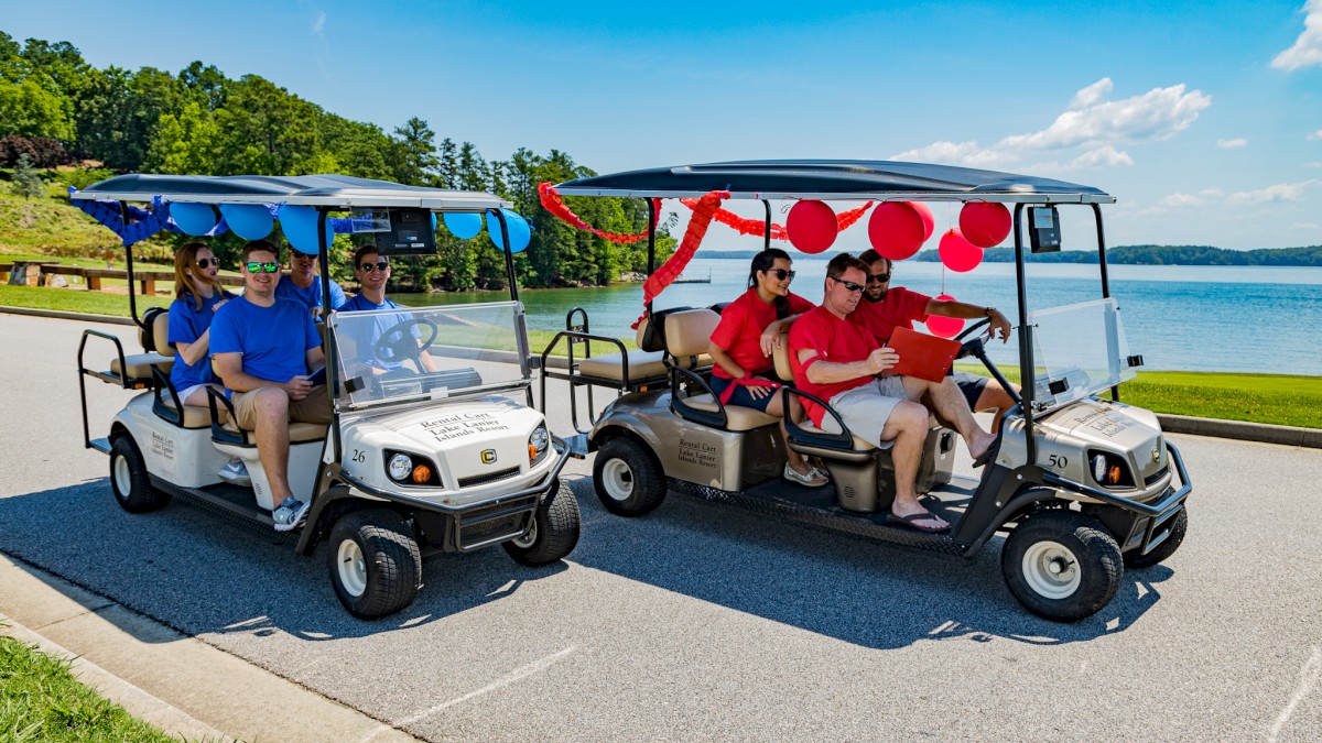 Two golf carts adorned with red and blue decorations, carrying people, are parked on a scenic road by the water under a clear sky.