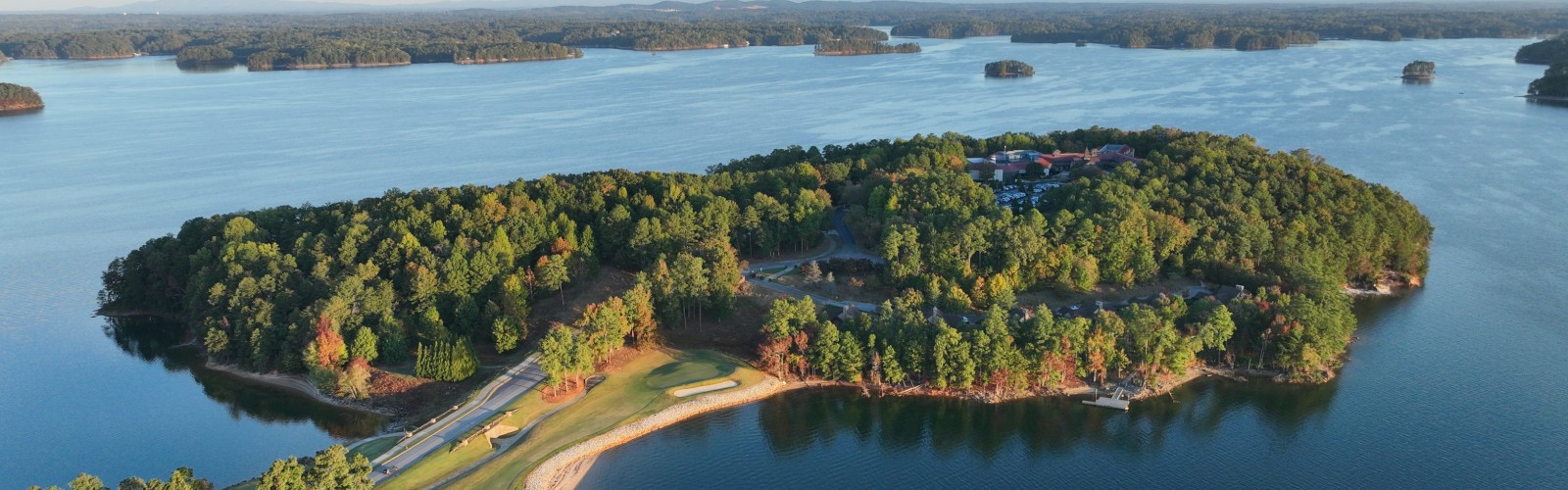 An aerial view of a lush, forested island surrounded by a calm lake, with winding pathways and clear skies in the background.