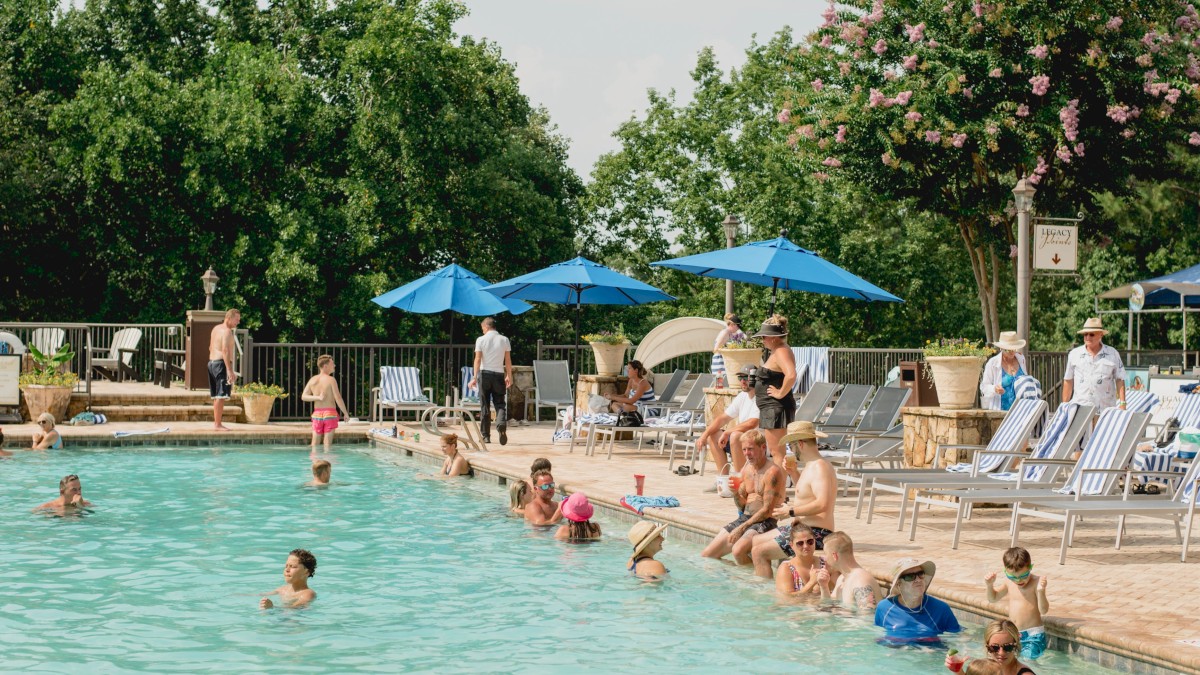 People are enjoying a sunny day at a swimming pool, with several lounging under blue umbrellas.