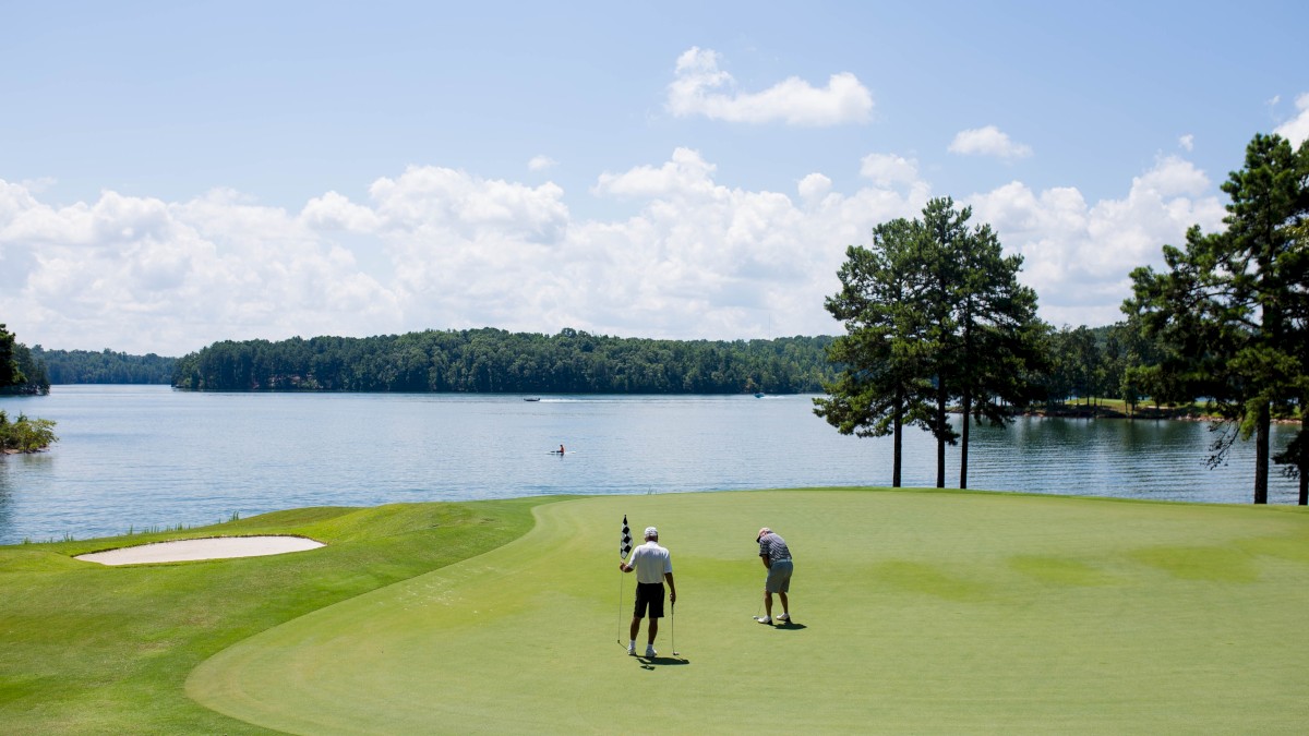 Two people are playing golf on a lush green course near a scenic lake, surrounded by trees under a partly cloudy sky.