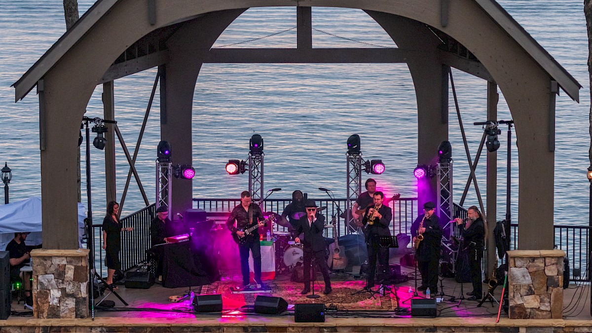 Outdoor concert by a lake with a band playing under a pavilion, people seated and standing, evening ambiance.