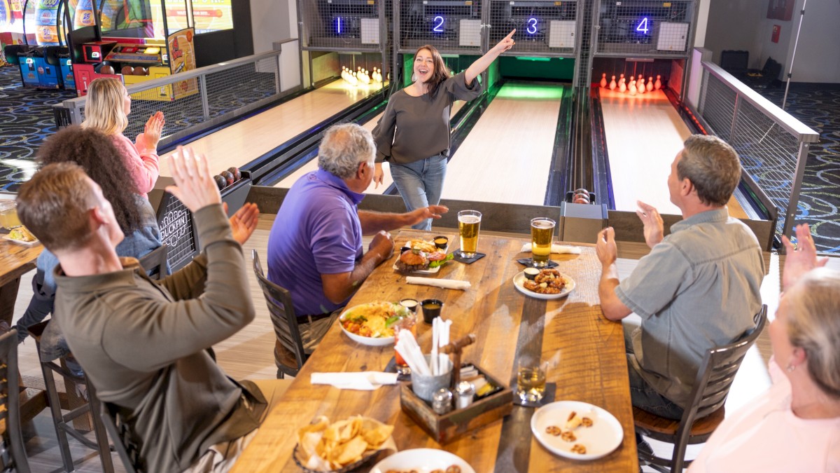 People are cheering a woman bowling as they enjoy food and drinks at a bowling alley.