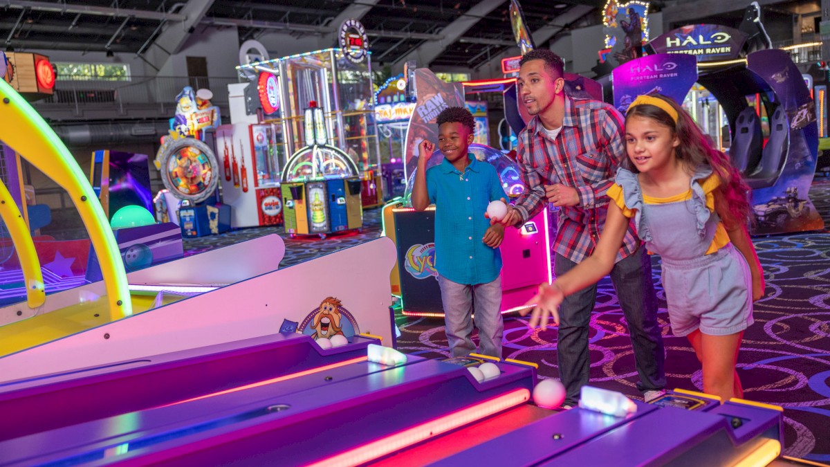 People playing skee-ball in a brightly lit arcade, surrounded by various game machines and neon lights.