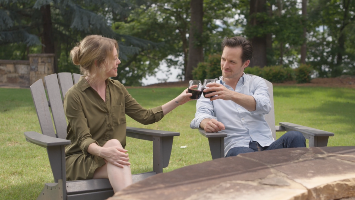 A couple sitting outside in wooden chairs, clinking glasses, with a green lawn and trees in the background.