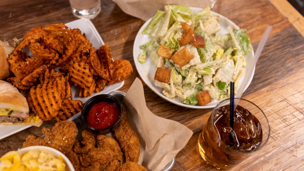 The image shows a table with fried chicken, waffle fries, a Caesar salad, mac and cheese, and a glass of iced beverage with a straw.