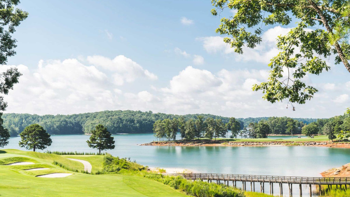 A scenic view of a lake surrounded by trees and a grassy golf course, with a bridge crossing over the water under a bright blue sky.