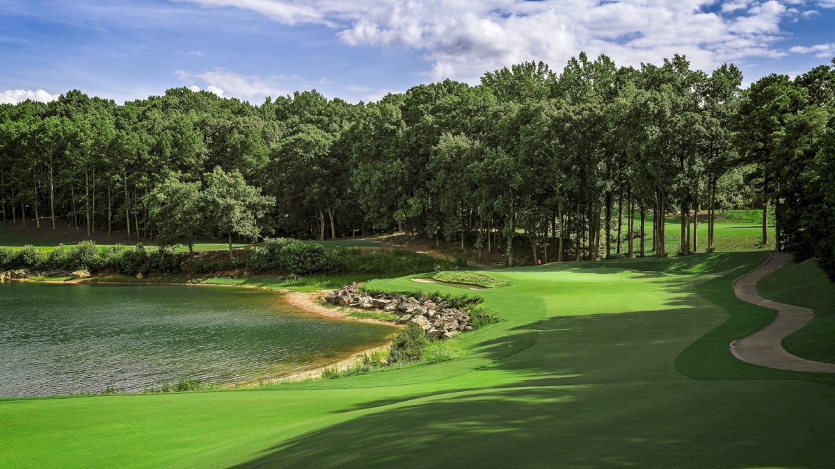 A scenic golf course with a water hazard, lush green grass, trees, and cloudy sky on a bright day.