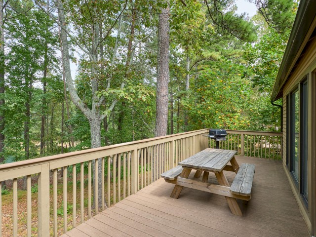 A wooden deck with a picnic table and grill overlooks a wooded area, adjacent to a house with large windows and a red door.