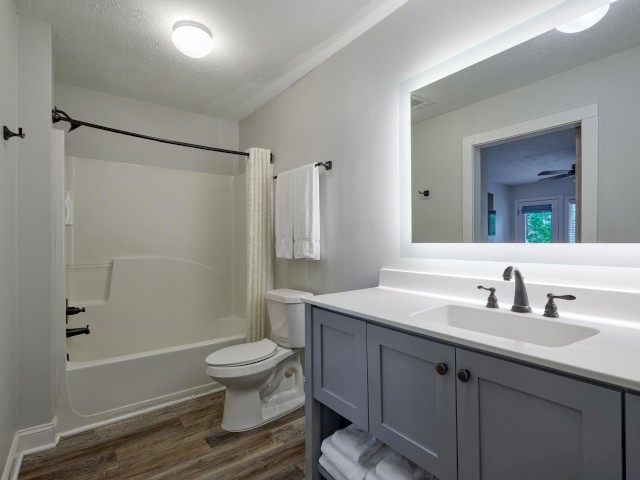 A modern bathroom with a bathtub, shower curtain, toilet, and sink. The mirror is lit, and towels are neatly stacked under the vanity.