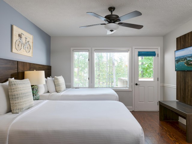 A hotel room with two beds, wall art, a ceiling fan, a TV, and large windows with a view of trees.