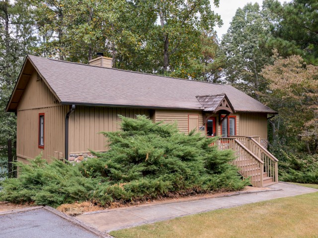 A small, rustic cabin with a porch and stairs surrounded by greenery and trees, featuring a sloped roof and wooden siding.